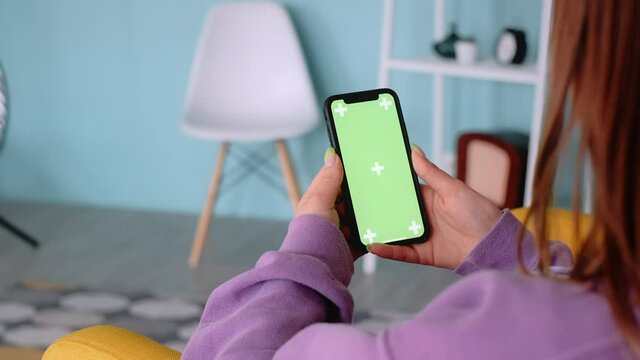 Young woman sitting on armchair at home and uses smartphone with green screen, tap motion. Mock-up. 4k	