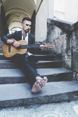 Male street musician playing the guitar outdoors with bare feet in old European city.