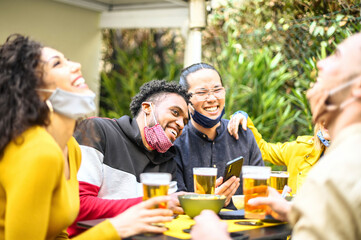 Multiracial people smiling with open face mask in the garden of restaurant while drinking beer - New normal lifestyle concept about happy friends having fun together at cocktail bar