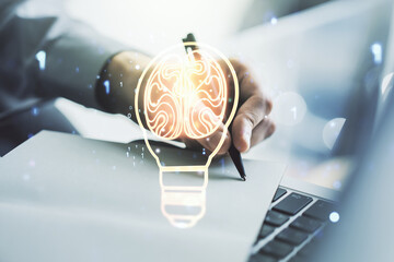 Creative light bulb with human brain hologram and hand writing in notepad on background with laptop, artificial Intelligence and neural networks concept. Multiexposure