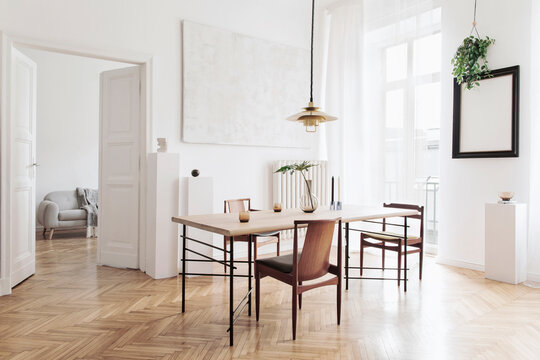 Stylish and modern dining room interior with design sharing table, chairs, gold pendant lamp, abstract paintings and elegant accessories. Tropical leafs in vase. Eclectic home decor.