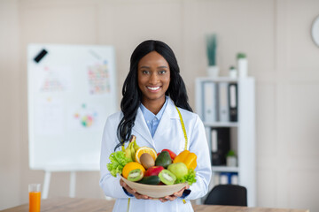 Portrait of happy black dietitian in lab coat holding bowl of fresh fruits and vegetables, smiling...