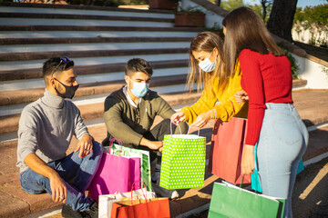 Group of young friends sitting on the steps of the city square wearing protective face masks against covid-19 coronavirus and look into their shopping bags at their newly made purchases