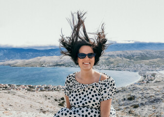 Smiling Caucasian woman in a polka dots flowy dress and sunglasses with hair blown up by the wind