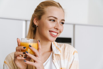 Joyful blonde woman drinking juice and smiling at home kitchen