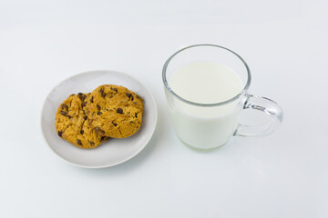 Two American cookies on white porcelain saucer and transparent cup of milk on white surface. Top view.