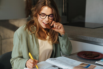 Pleased nice student woman eating cookie while doing homework at home