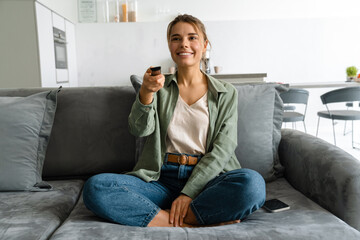 Obraz premium Happy nice woman watching TV with remote control while sitting on sofa