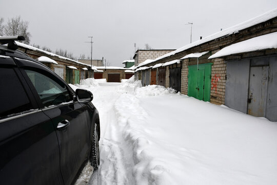 A cloudy winter day. Snowfall. The car is parked between one-story brick garages with closed metal painted gates. Drifts of snow near the walls along the road. Snow on the roofs.