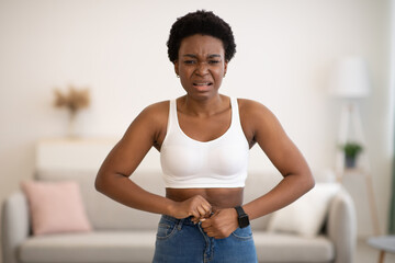 Frustrated Black Woman Buttoning Small Jeans After Weight Gain Indoors