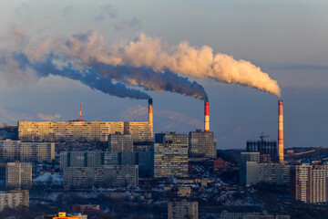 The smoking chimneys of TPP-2 in Vladivostok amid residential buildings and distant snowy mountains. Smoke in the city.