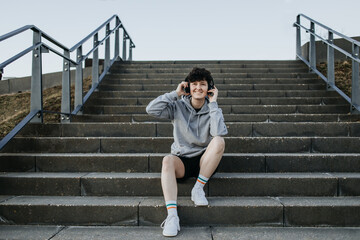 Junger Mensch sitzt auf Treppe und hört Musik mit Kopfhörern sportlich gekleidet 