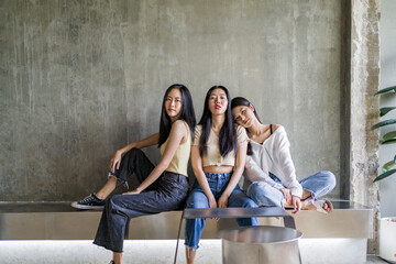 Three happy young Asian female friends casually pose sitting together inside a rustic room against bare concrete wall