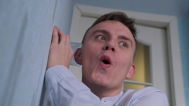 Emotional young man eavesdrops on neighbors putting his ear to the wall