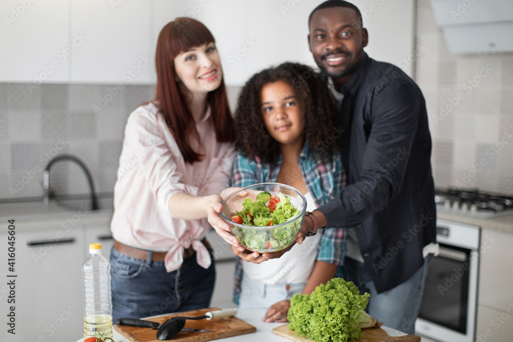 Wall mural Multiethnical family, smiling and demonstrating fresh vegetable salad to the camera. Family cooking in the kitchen at home