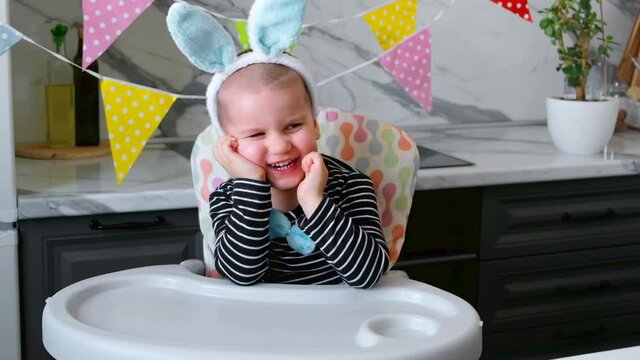 Funny Blond Boy Kid with Rabbit Ears plays with colored Easter Eggs sitting at the Kitchen table. Holidays Concept.
