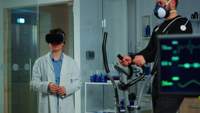 Scientist doctor in sport science laboratory wearing virtual reality goggles while athlete running with electrodes attached on body monitoring physical endurancewhile ekg scan runs on computer screen.