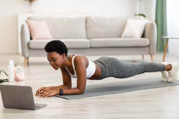 African Lady Standing In Plank Position Exercising At Laptop Indoor