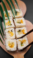 Dessert sushi. Sweet kiwi, pineapple sushi rolls. Sushi on a wooden tray on black background with tropical leaf. Vertical photo. Holding a sweet roll