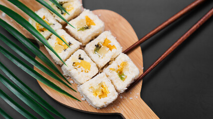 Dessert sushi. Sweet kiwi, pineapple sushi rolls. Sushi on a wooden tray on a black background. Holding a sweet roll with wooden sticks.