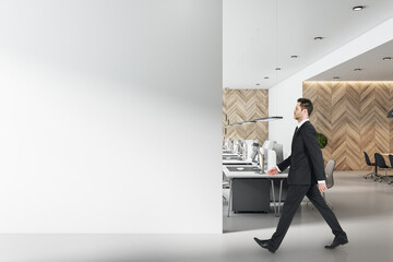 Businessman in black suit goes by modern workplaces in sunny open space office with blank light...