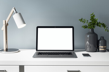 Front view on blank white laptop display on white table with stylish vase, lamp and smartphone. Mock up