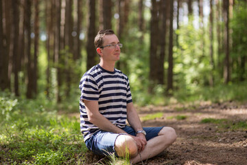 Naklejka na ściany i meble A man with glasses sits on the ground in a bright sunny spring forest.