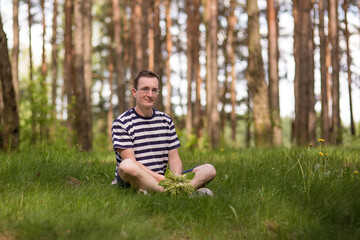Naklejka na ściany i meble Portrait of a smiling man in glasses with a bouquet of fresh lilies of the valley. He sits on the grass in a bright sunny spring forest.