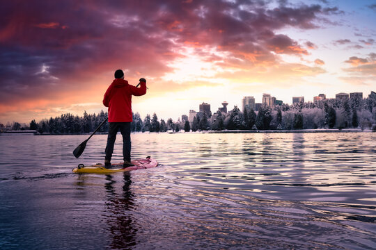 Adventurous male is paddle boarding near Stanley Park with Downtown City Skyline in the background. Taken in Vancouver, BC, Canada. Colorful Sunrise Sky Art Render.