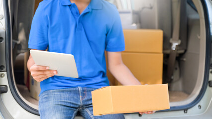 Delivery man sit at the car uses a tablet and holds a parcel box to deliver the goods to the...