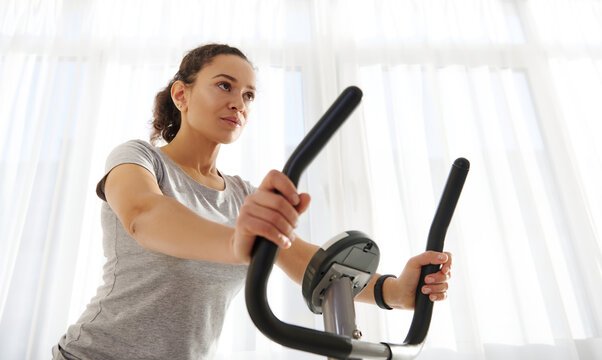 Attractive Fit Woman Exercising On A Spin Bike At Home On A Beautiful Sunny Day. Cardio Workout. Fitness At Home. Closeup