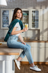 joyful woman looking at camera while sitting on kitchen table with glass of fresh juice