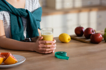cropped view of woman near glass of orange juice, fresh fruits and measuring tape in kitchen