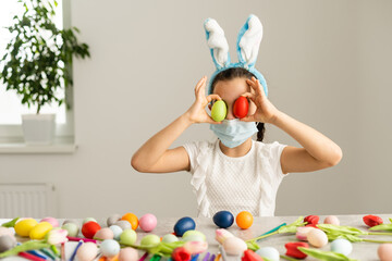 Coronavirus and Easter holiday. Cute little girl in a protective medical mask with bunny ears with Easter eggs and spring flower.