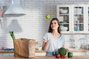 smiling woman juggling with ripe apple near paper bag and fresh vegetables in kitchen