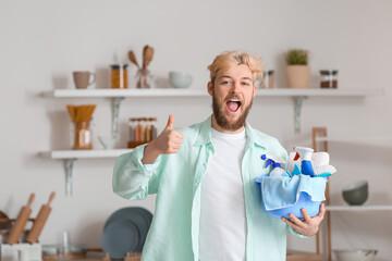 Young man with cleaning supplies showing thumb-up in kitchen
