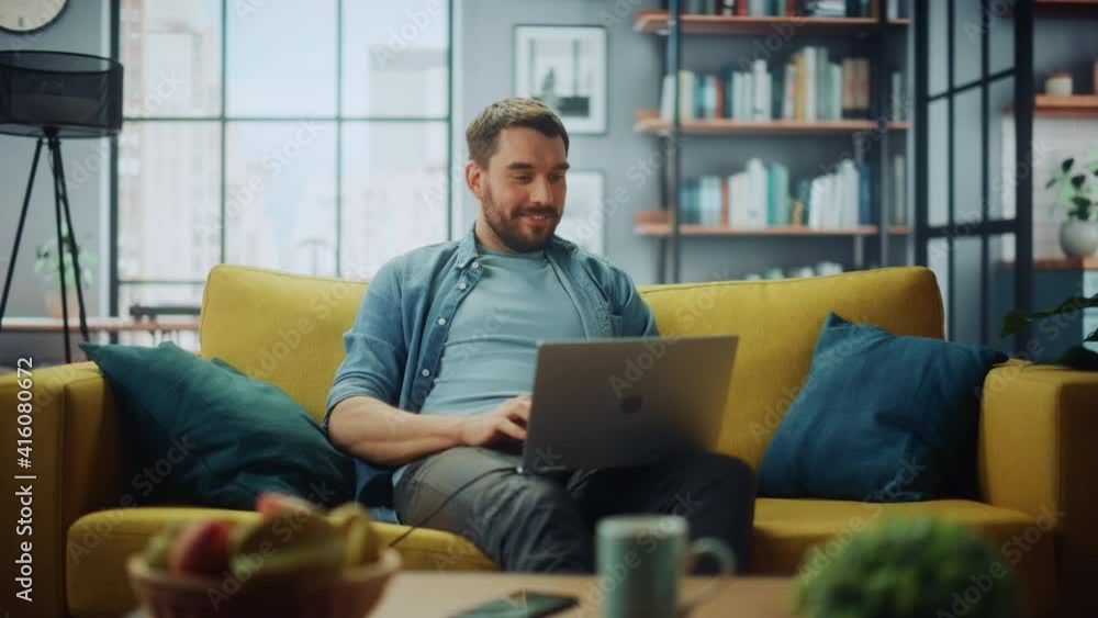 Poster handsome caucasian man working on laptop computer while sitting on a sofa couch in stylish cozy livi