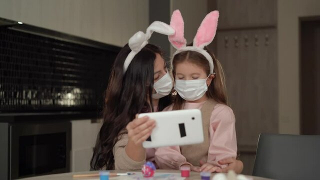 Happy Easter. A cheerful family is preparing for the Easter holiday. Mom and daughter dressed in medical masks are sitting at home in the kitchen. make a video call via a tablet