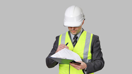 Man in suit and reflective vest with white hardhat on his head, looks to sheets of paper on...