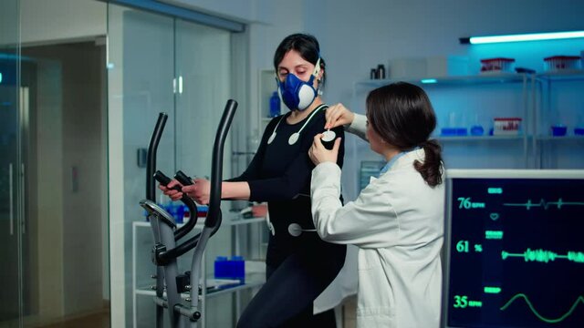 Woman athlete with mask running on cross trainer after medical researcher attached electrodes to her body. Physician using computer to controls EKG data showing on laboratory monitors, VO2 max test