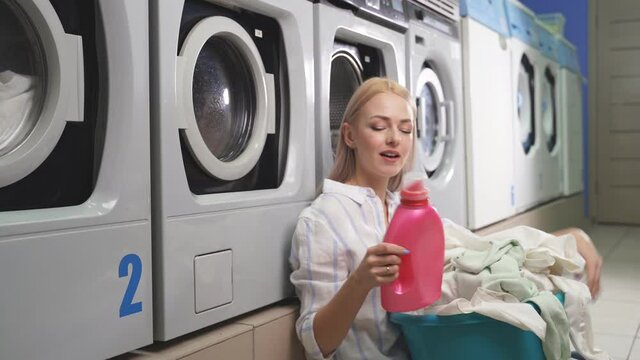 Portrait of an attractive blonde woman in a public laundry room next to a washing machine, holding a basin of laundry and smiling at the camera.
