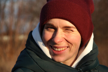 
A blue-eyed girl in a burgundy knitted hat and a white hood smiles, winking with one eye against...
