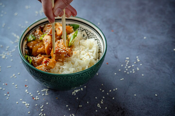 Woman eating with chopsticks traditional Asian street food - Crispy sesame chicken in sweet and sour sauce with portion of rice in a green decorated bowl. Homemade dish, ready to eat. Gray background © Uniqueton