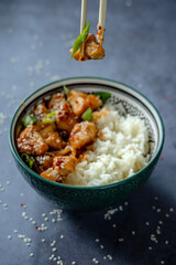 Hand eating sweet and sour sauce chicken using chopsticks. Traditional Asian street food - Crispy sesame chicken with rice in a green decorated bowl. Homemade dish, ready to eat. Gray background