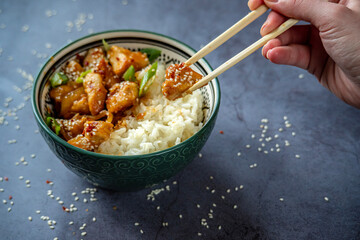 Woman eating with chopsticks traditional Asian street food - Crispy sesame chicken in sweet and sour sauce with portion of rice in a green decorated bowl. Homemade dish, ready to eat. Gray background