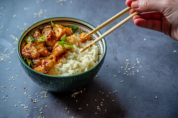 Hand eating with chopsticks traditional Asian street food - Crispy sesame chicken in sweet and sour sauce with portion of rice in a green decorated bowl. Homemade dish, ready to eat. Gray background