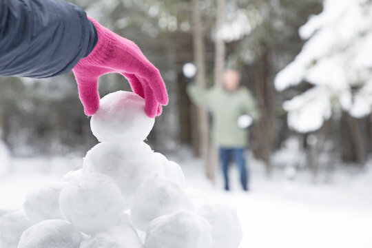 Original winter photograph of a woman's pink gloved hand picking up a snowball from a snowball pile for to have a snowball fight with man in the distance.