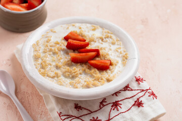 Simple oatmeal porridge with strawberries in a white plate on a linen napkin on pink background. Breakfast health food concept. Top view