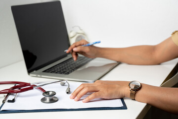 a doctor working on a laptop taking notes at the workplace. A female physician writing in a notebook using computer sitting at desk.