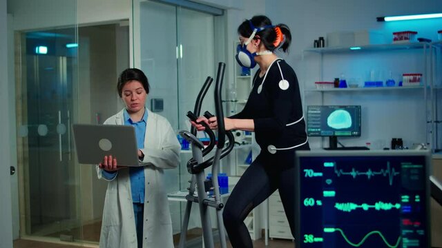 Doctor scientist holding laptop computer and controling EKG data showing on laboratory monitors. Lab science researcher measuring endurance of athlete, heart rate using electrodes and mask